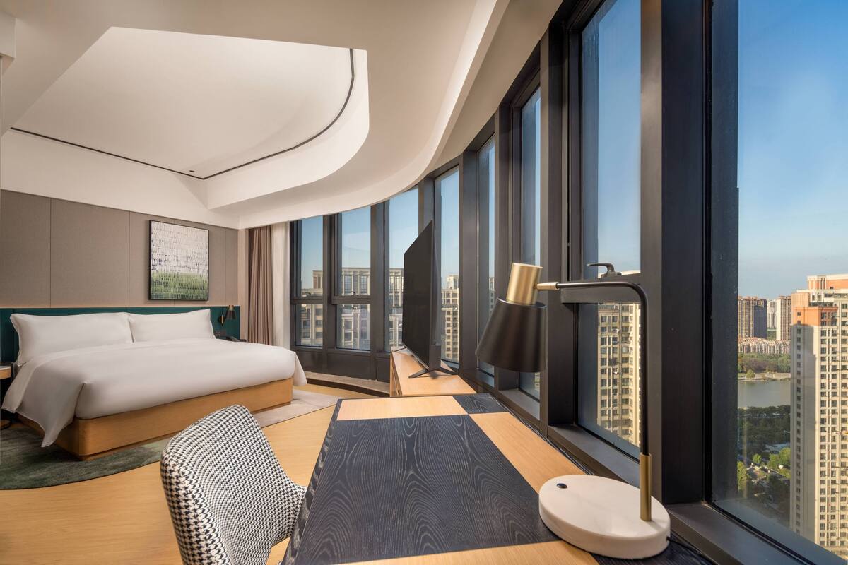 A modern hotel room with a large bed, a desk, and a view of the cityscape through large windows.