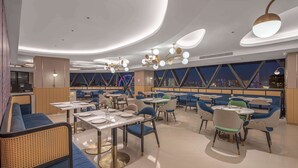 Un restaurante moderno con mesas redondas, sillas azules y verdes, y vistas a la ciudad a través de amplios ventanales.