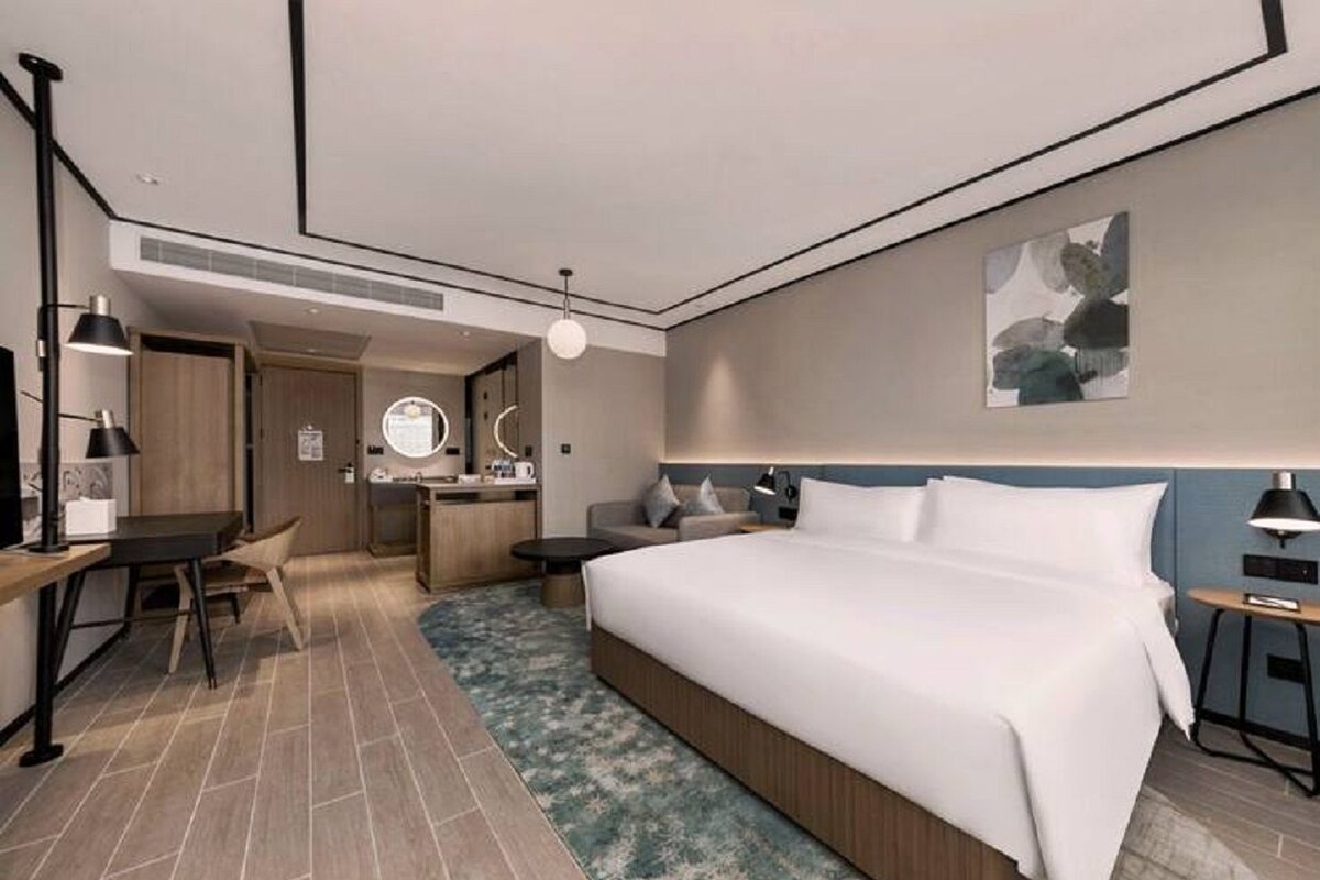 Una habitación de hotel moderna con una cama grande, un escritorio, una silla, una mesita pequeña, un espejo redondo y una lámpara de pared.