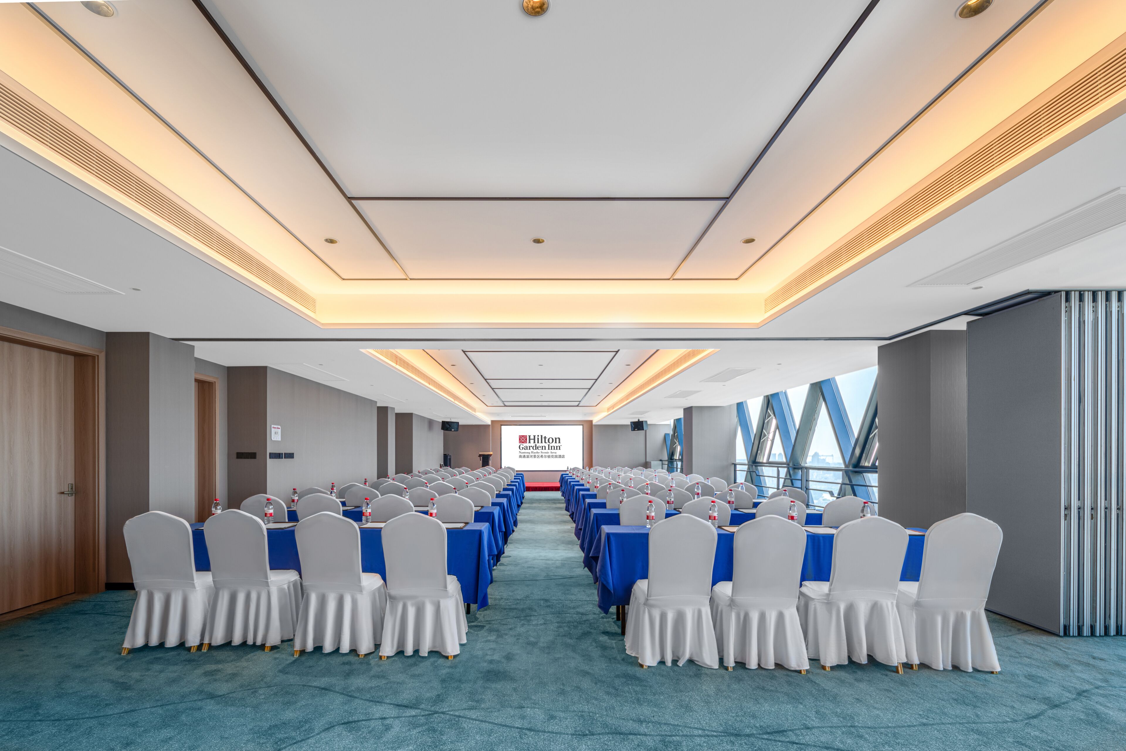 A conference room with rows of tables and chairs, a large screen at the front, and a view of the city through large windows.