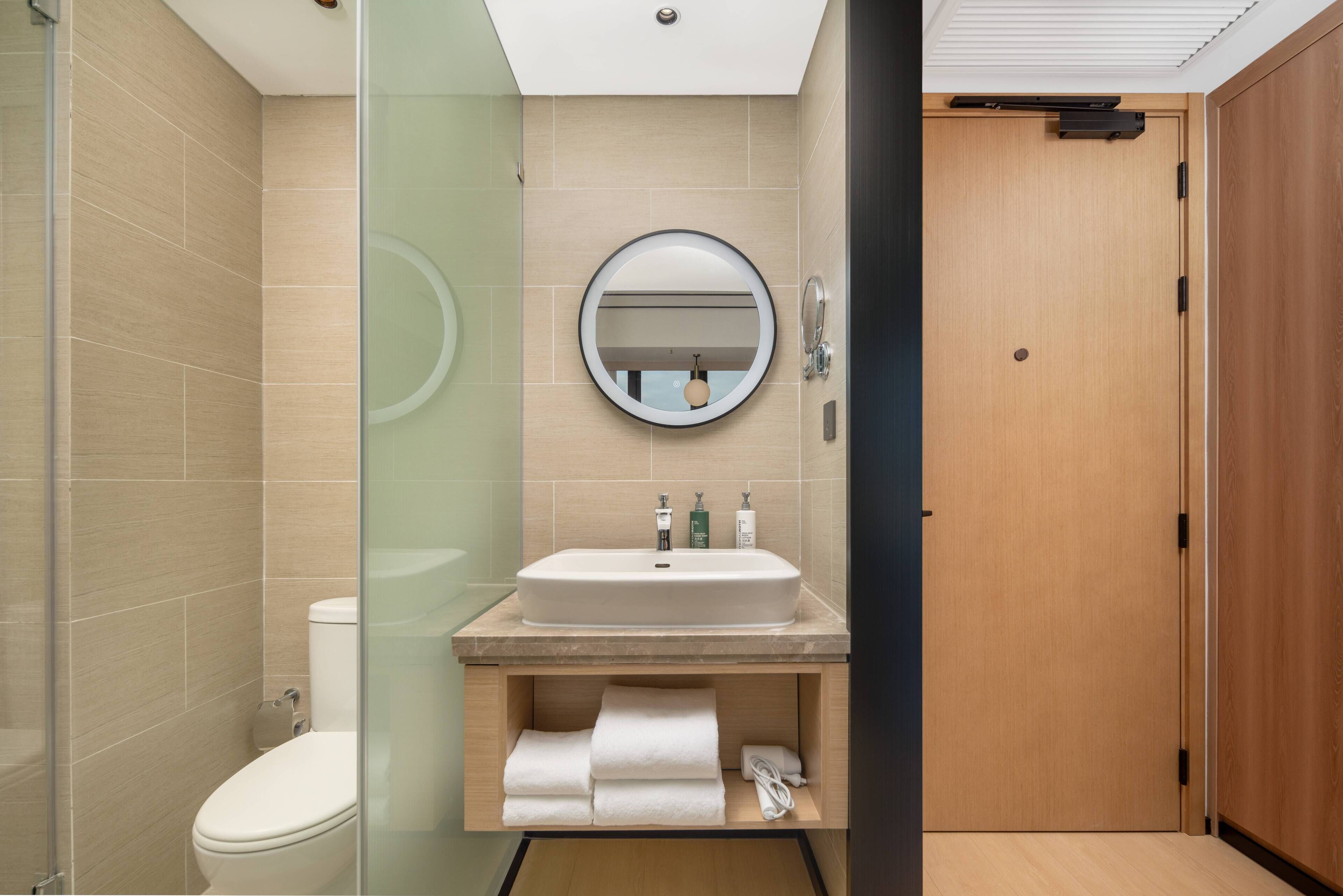 Un baño moderno con cabina de ducha de vidrio, un lavamanos blanco con espejo redondo y una puerta de madera.
