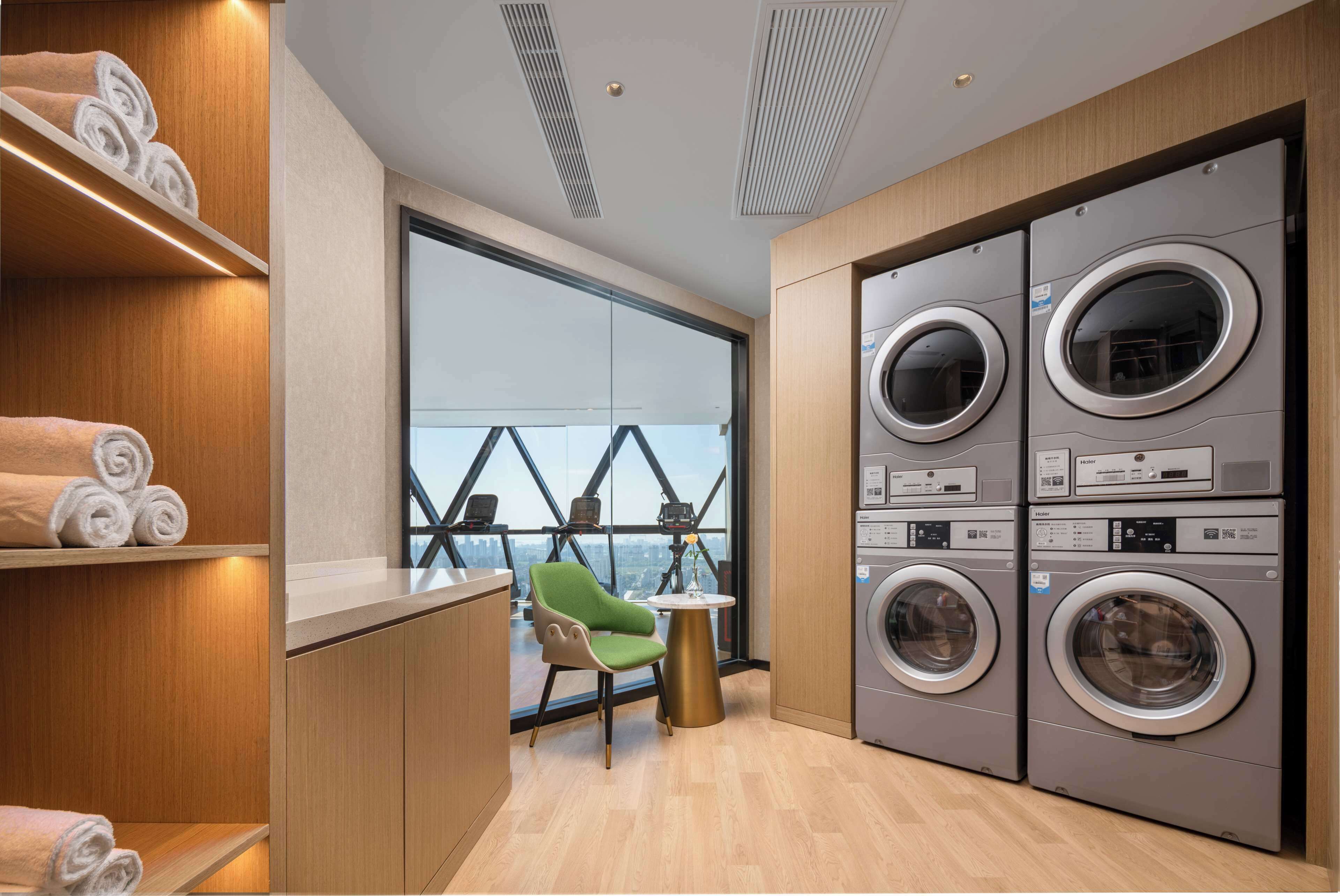 A modern laundry room with stacked washing machines, a green chair, and a view through large windows.