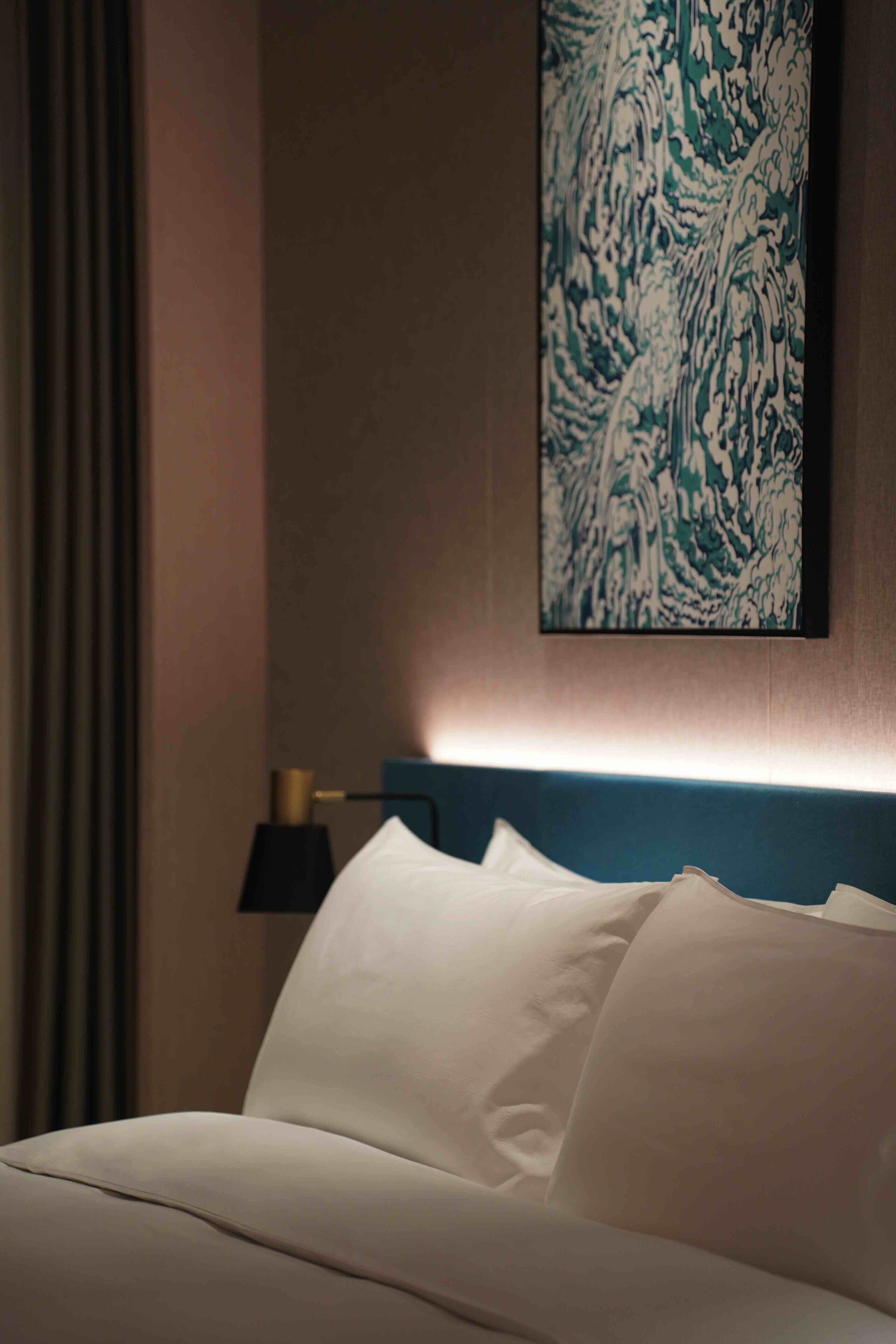 A neatly made bed with white linens, a wall-mounted abstract art piece, and a bedside lamp.
