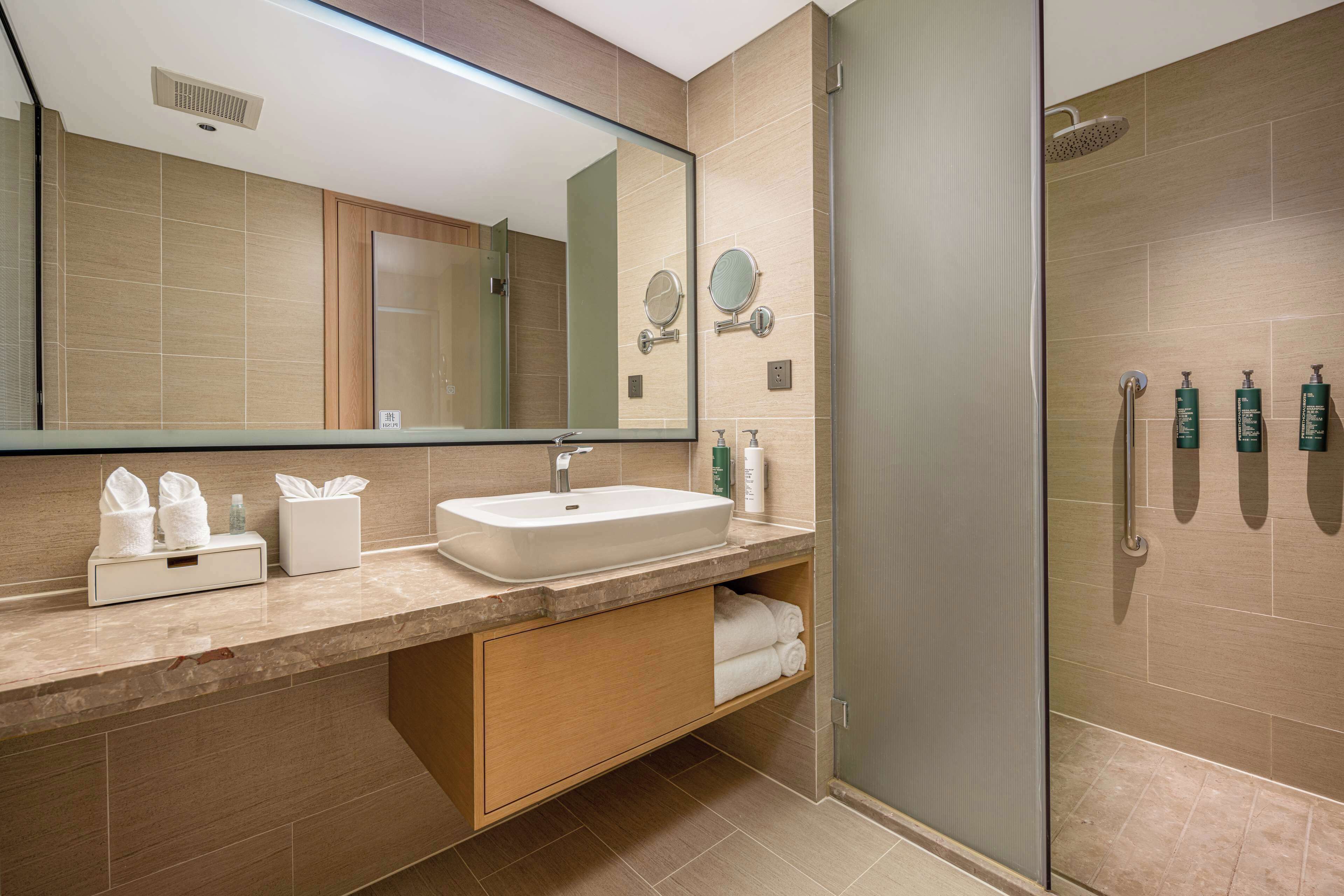 A modern bathroom with a large mirror, a sink, a glass shower enclosure, and toiletries on the vanity.