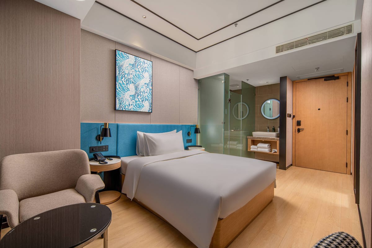 A modern hotel room with a large bed, a seating area, a bedside table, a desk, a mirror, and a bathroom with a sink and mirror.