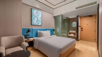 Una habitación de hotel moderna con una cama grande, una zona de estar, una mesita de noche, un escritorio, un espejo y un baño con lavabo y espejo.