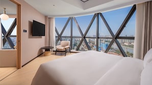 Habitación de hotel moderna con una cama grande, un televisor montado en la pared, una silla, una mesita y un ventanal con vistas a la ciudad.