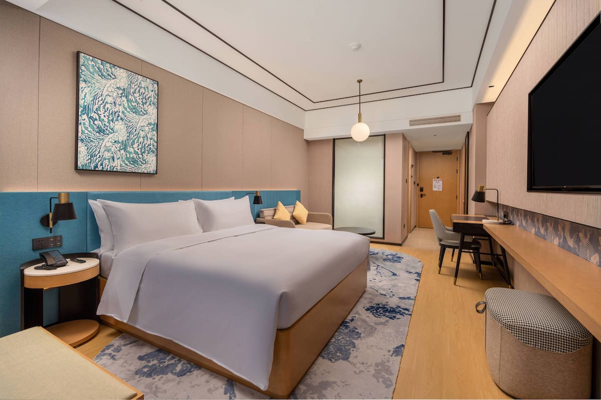 A modern hotel room with a large bed, a desk, and a chair. There is a painting on the wall, a lamp, and a trash bin.