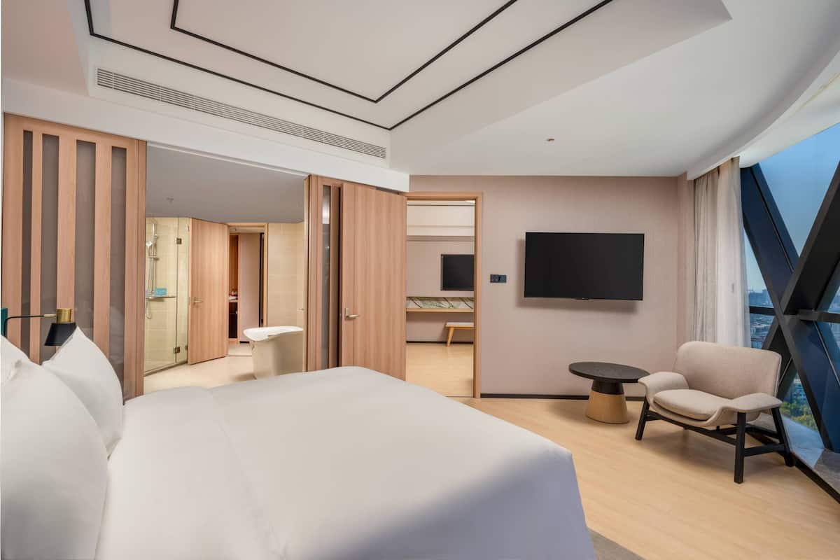 A modern hotel room with a large bed, a seating area with a table, a TV mounted on the wall, and a view of the city through large windows.