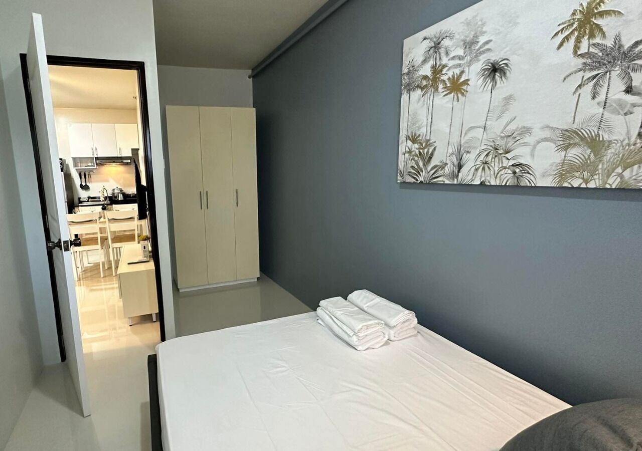 A bedroom with a bed, a painting of palm trees, and a view into the kitchen.