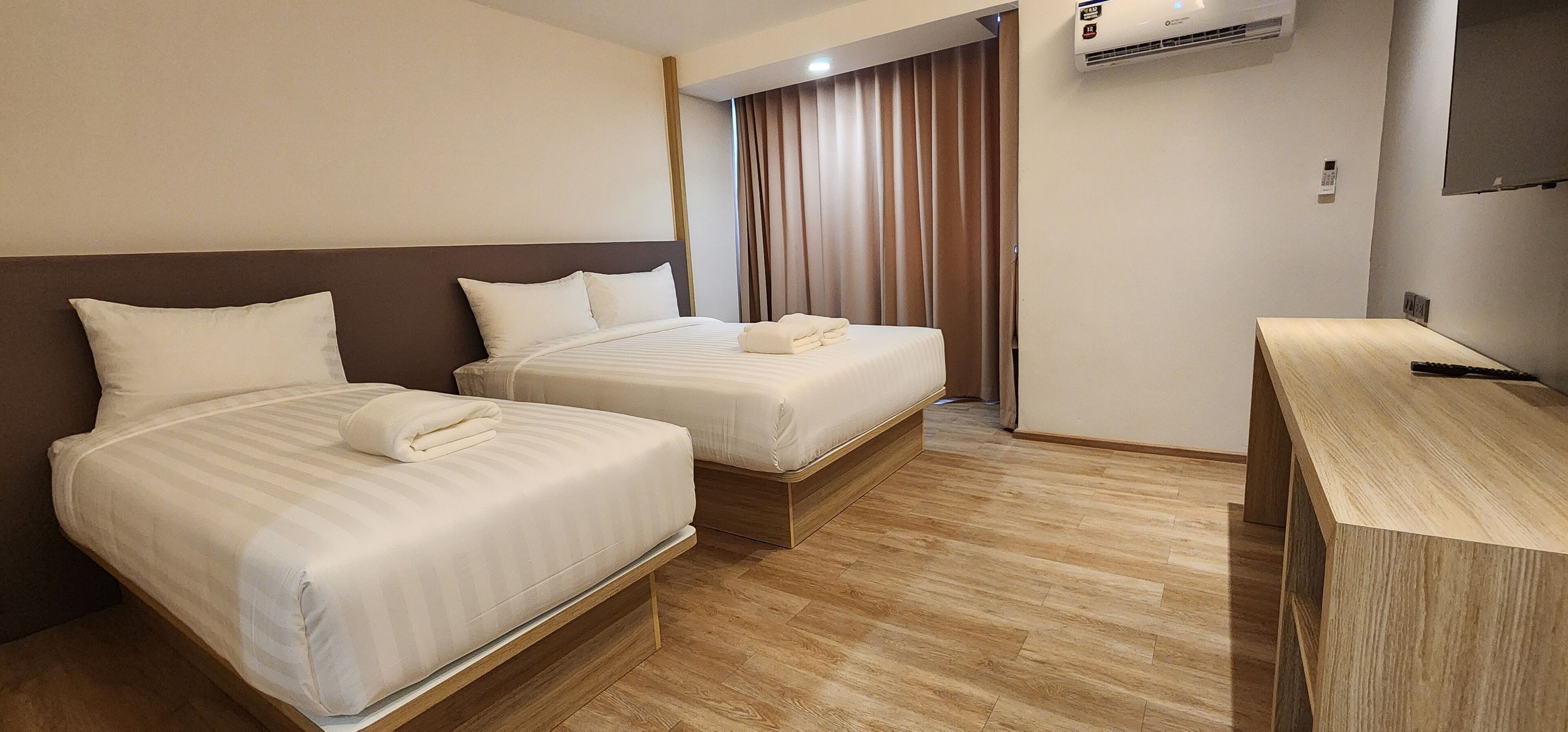 A hotel room with two beds, a wooden desk, an air conditioning unit, and a curtain.