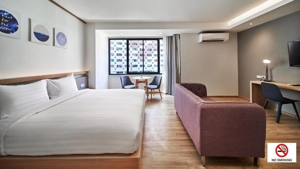 A hotel room with a bed, a desk, a chair, a sofa, and a TV. There is a window with a view of buildings outside. - Prom Thailand Cultural Centre Hotel Annex2 (Bangkok)