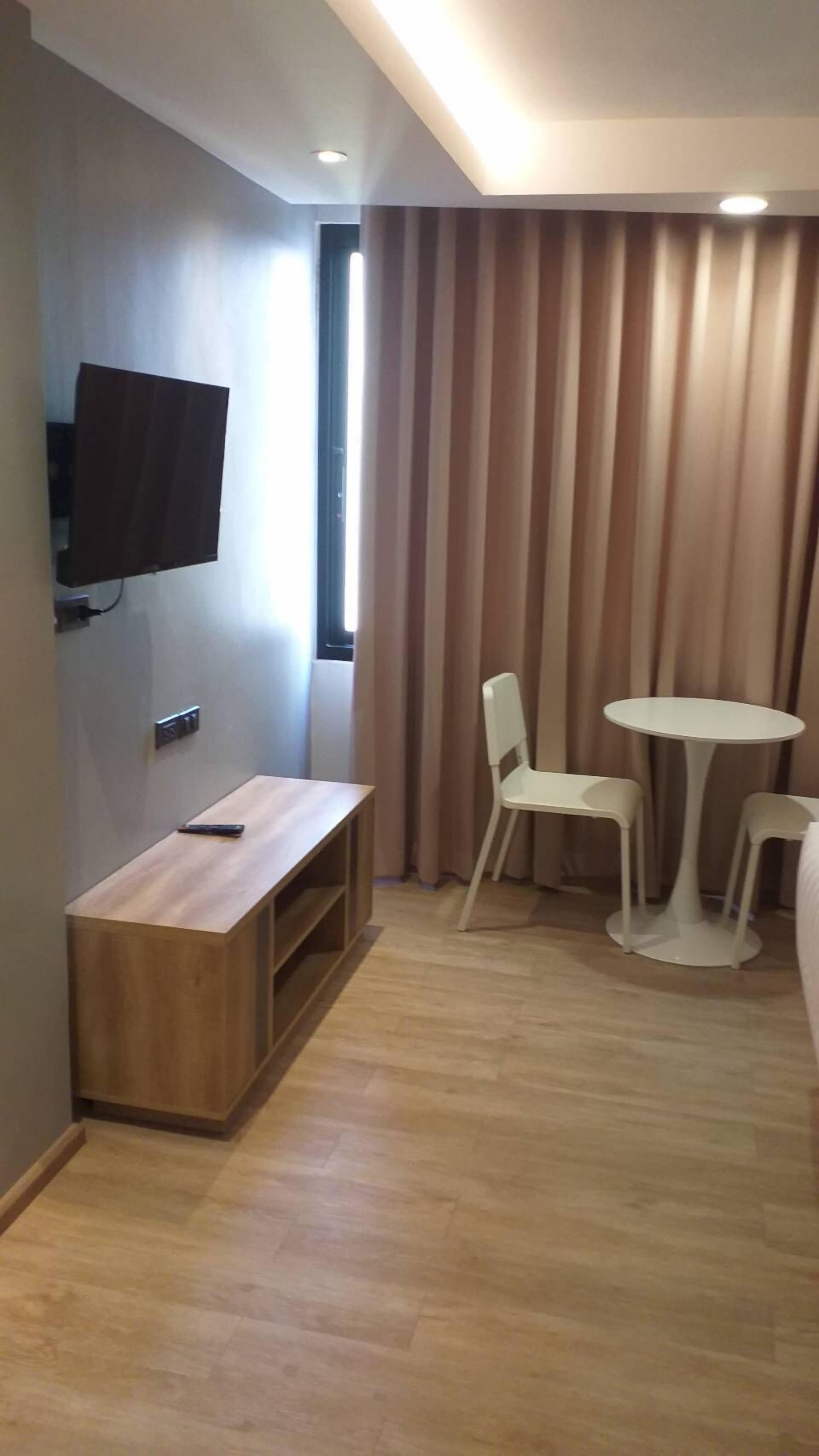 A modern room with a wooden bench, a TV mounted on the wall, and a window with curtains. There are two white chairs and a small round table.