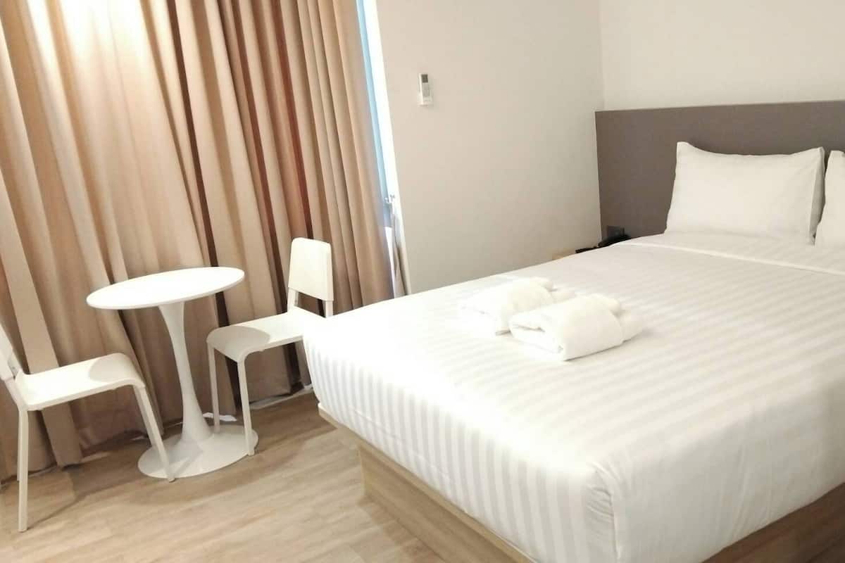Une chambre d’hôtel avec un grand lit, deux chaises blanches, une petite table ronde et une fenêtre avec des rideaux.