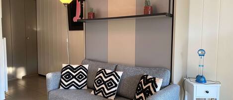 A modern living room with a grey sofa, patterned cushions, a floor lamp, a small table with a lamp, and a potted plant on a shelf.