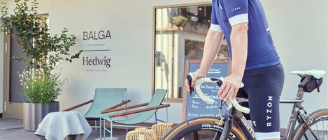 Un ciclista con camisa azul y pantalon corto negro, con una bicicleta estacionada frente a un edificio que tiene un letrero que dice "BALGA Hedwig".