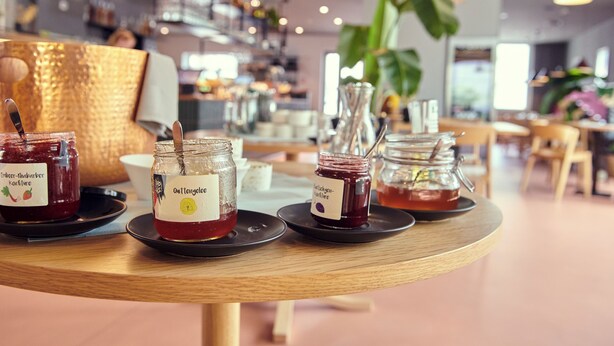 A table with jars of jam and honey, a woven basket, and a coffee machine in a cafe setting. - Hotel Balga Natur- und Bikehotel (Balgach)