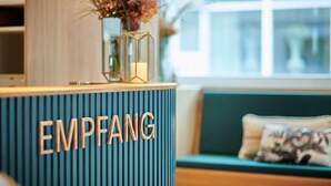A reception desk with the word "EMPFANG" on it, a vase with flowers, and a window in the background. - Hotel Balga Natur- und Bikehotel (Balgach)