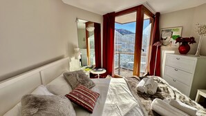 A hotel room with a large bed, a small table, a dresser, and a view of a snowy landscape through a window. - Luxury 4beds ski-in&out steps from center     (cervinia)