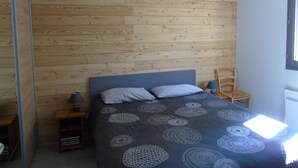A bedroom with a wooden wall, a bed with a patterned bedspread, a nightstand with a lamp, and a chair.