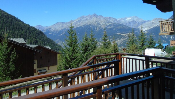 A wooden cabin with a mountain view, a wooden deck, and a white van in the foreground.