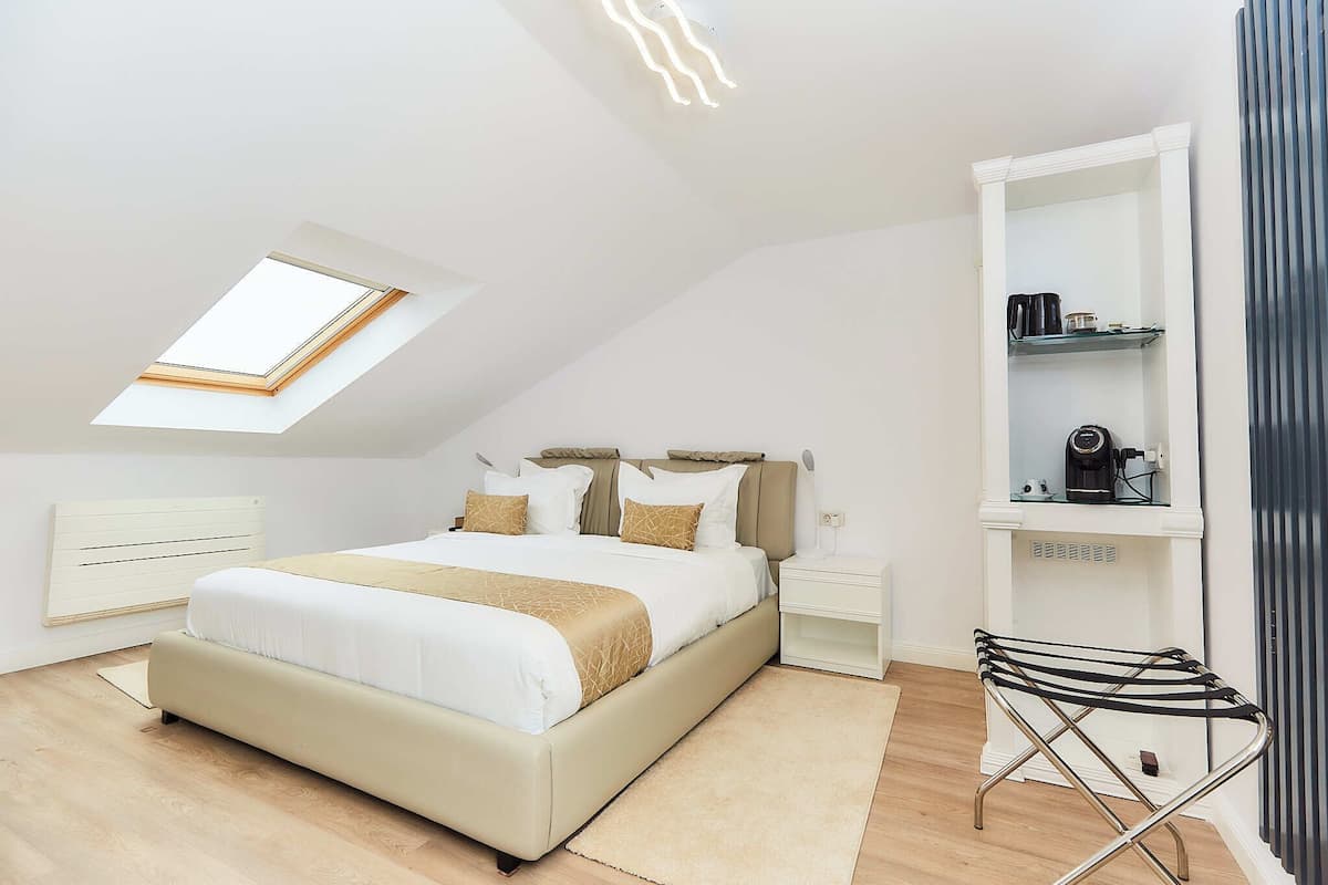 A modern bedroom with a skylight, a bed with beige bedding, a bedside table, a nightstand, a radiator, and a shelf with a kettle.