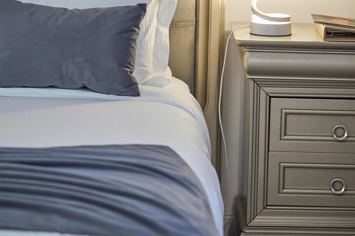 A neatly made bed with a headboard, a bedside lamp, and a nightstand with drawers.