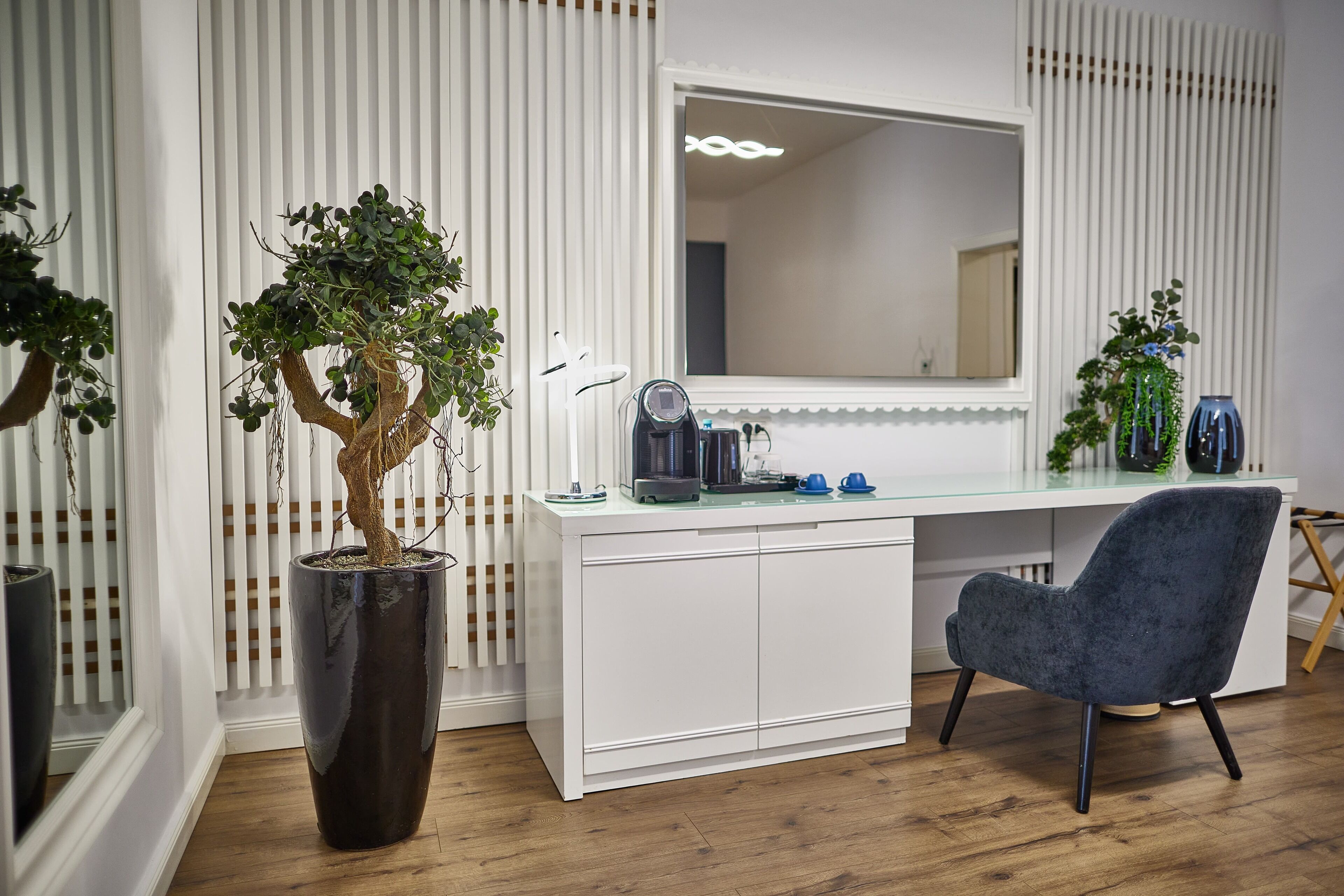 A modern room with a white desk, a large mirror, a potted plant, and a grey chair.