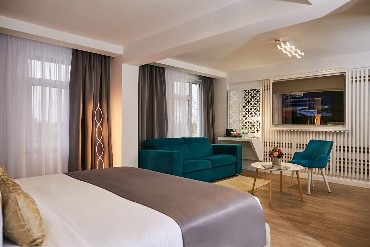 A modern hotel room with a large bed, a teal sofa, a small round table, and a blue chair.