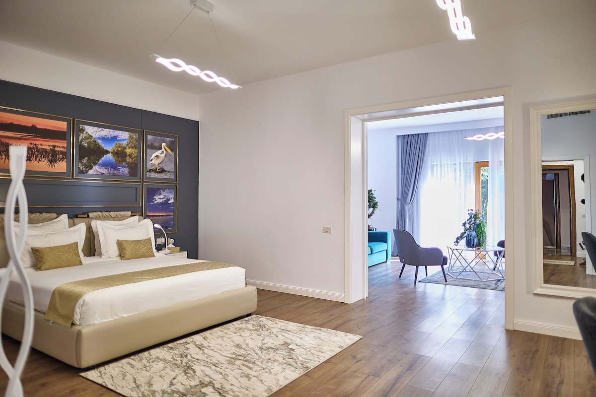 A modern bedroom with a large bed, a headboard with artwork, a bedside table, and a view into the living area.