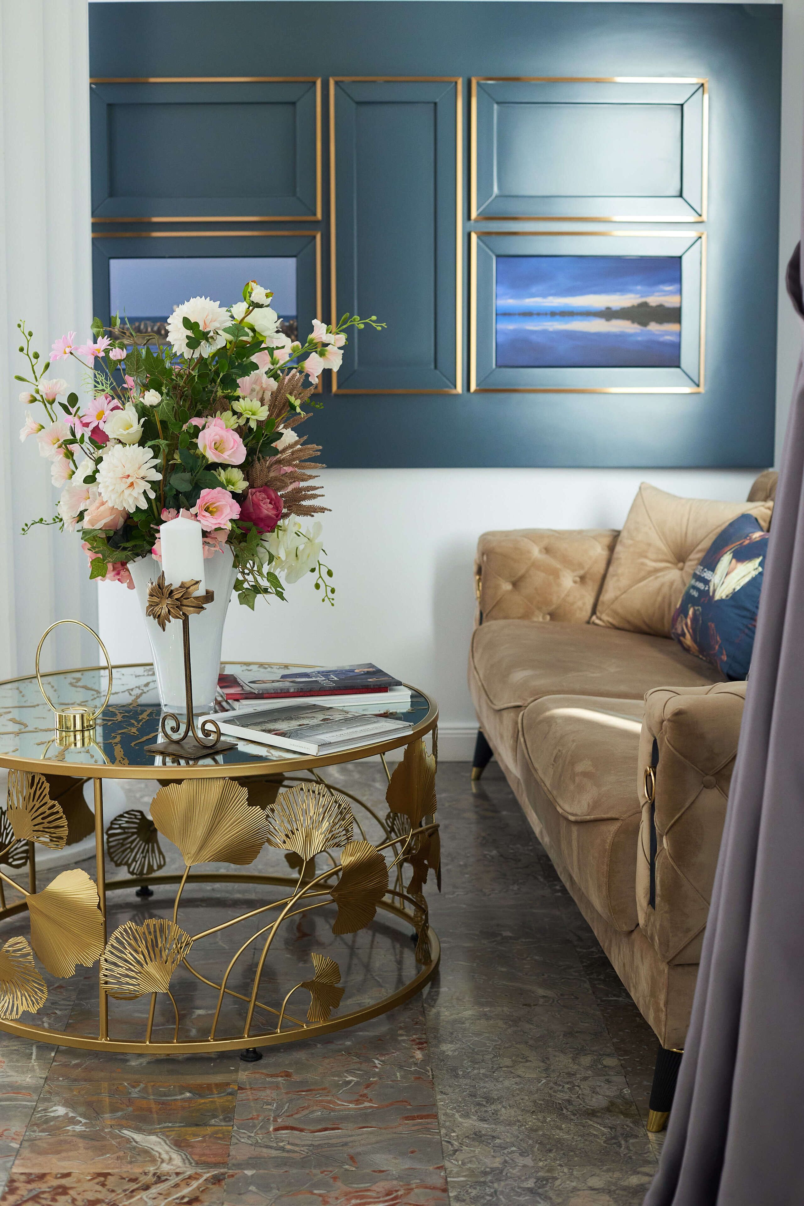 Un salón con un sofá color beige, una mesa de centro redonda de cristal con base dorada, y un jarrón blanco con flores.