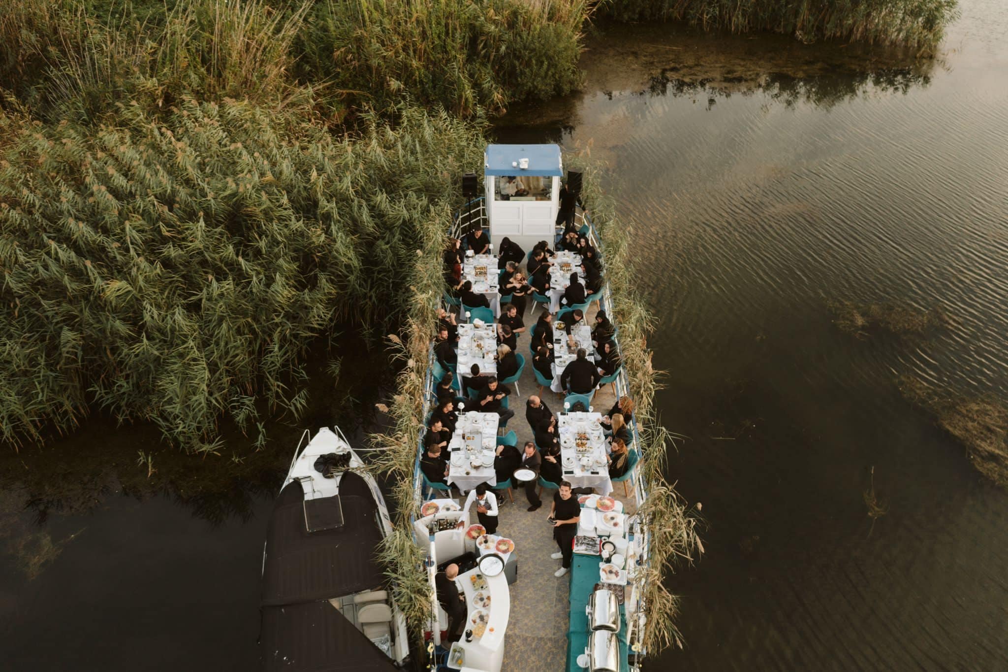 Vista aérea de um restaurante em barco com mesas e cadeiras dispostas ao longo das laterais, cercado por água e vegetação densa.