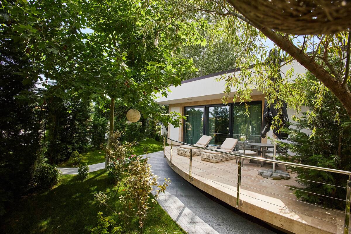 A modern outdoor deck with lounge chairs and a dining area, surrounded by lush greenery and trees.
