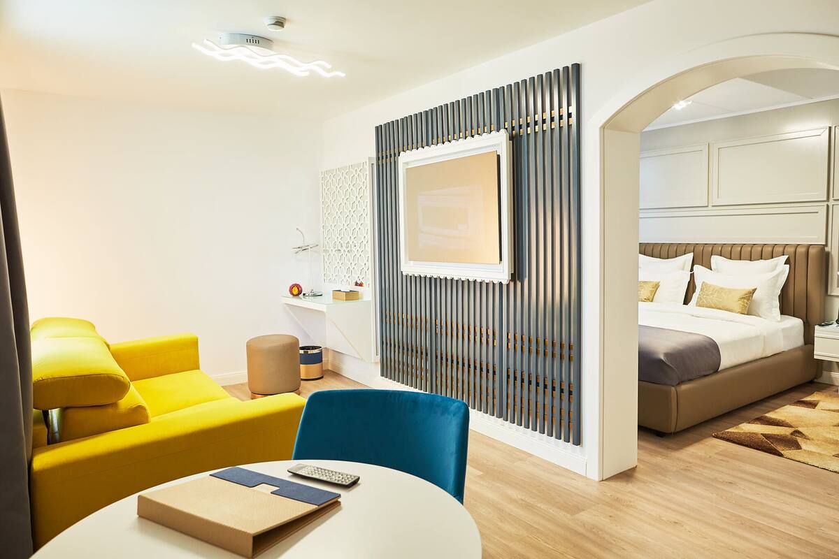 A hotel room with a yellow sofa, a blue chair, a round table with a remote control, a bed with a headboard, and a decorative wall panel.