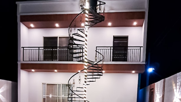 A modern building with a spiral staircase leading to a balcony, a swimming pool, and a covered outdoor area. - Roomshala 135 Paradise Villa Banglore (Devanahalli)