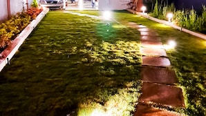 A well-lit residential area with a paved pathway leading to a white building with a balcony. - Roomshala 135 Paradise Villa Banglore (Devanahalli)