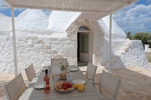Une table dressée à l’extérieur, recouverte d’une nappe blanche, avec des assiettes, une bouteille et un saladier rempli de fruits.