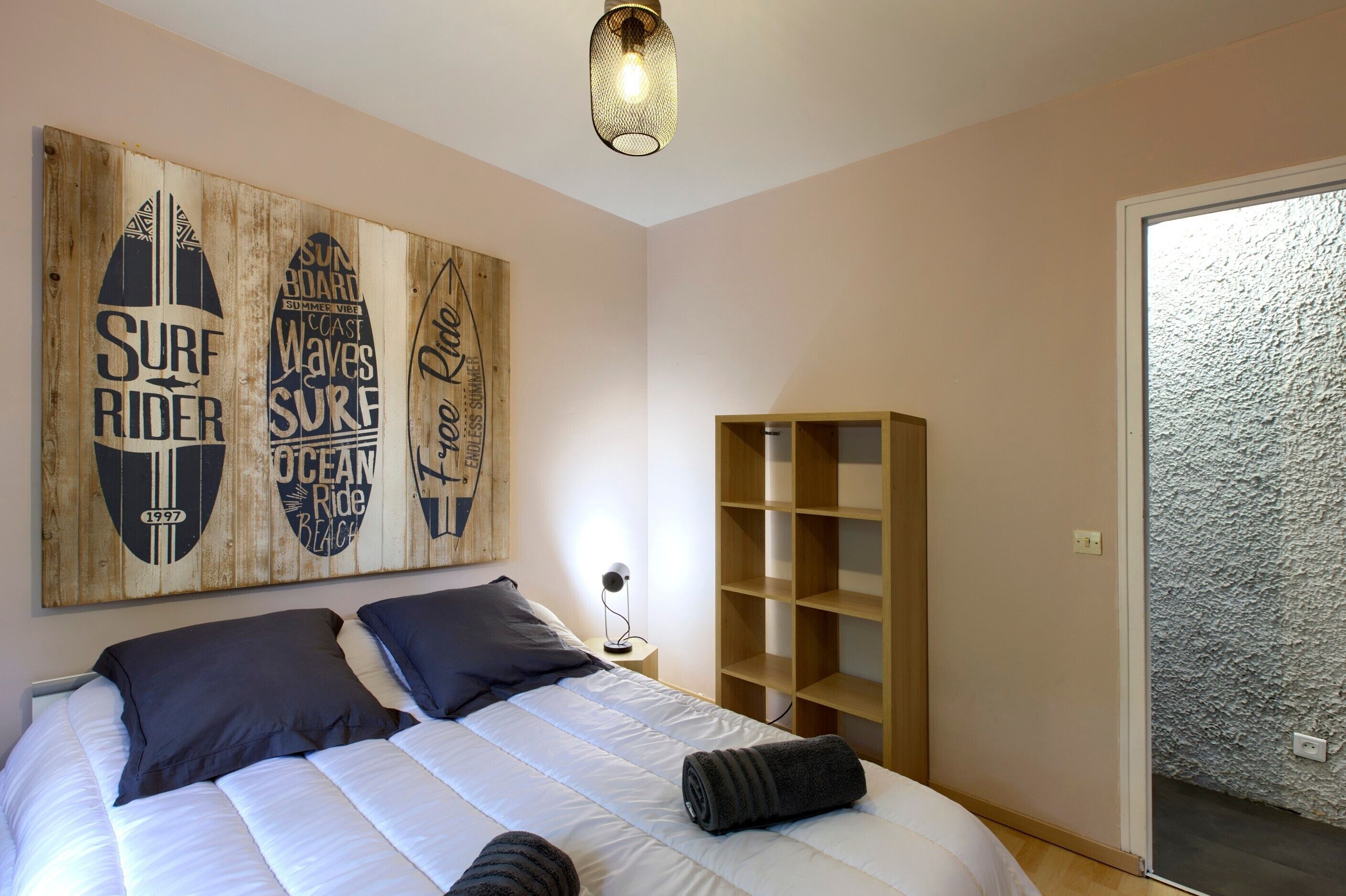 Une chambre avec un lit, des oreillers, une table de chevet, une lampe, une étagère en bois et des œuvres d’art sur le thème du surf accrochées au mur.