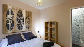 Une chambre avec un lit, des oreillers, une table de chevet, une lampe, une étagère en bois et des œuvres d’art sur le thème du surf accrochées au mur.