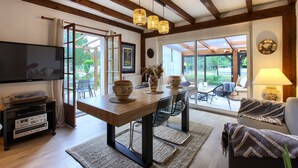 A dining area with a wooden table, chairs, a television, and a view through large windows. - Villa 'Rodolphe' with Private Pool, Private Terrace, and Wi-Fi (Le Pian-Médoc)