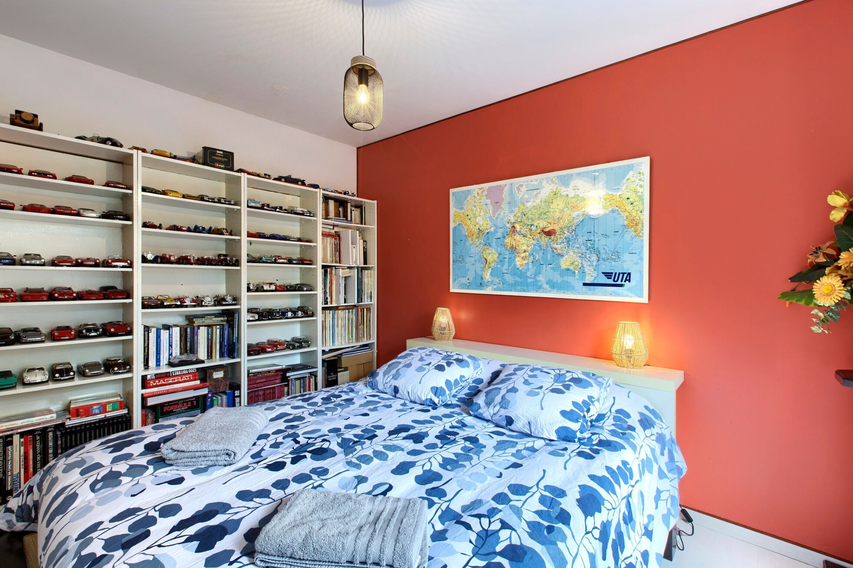 Un dormitorio con una cama, una estantería con maquetas de coches, un mapa mundial y un jarrón con flores.