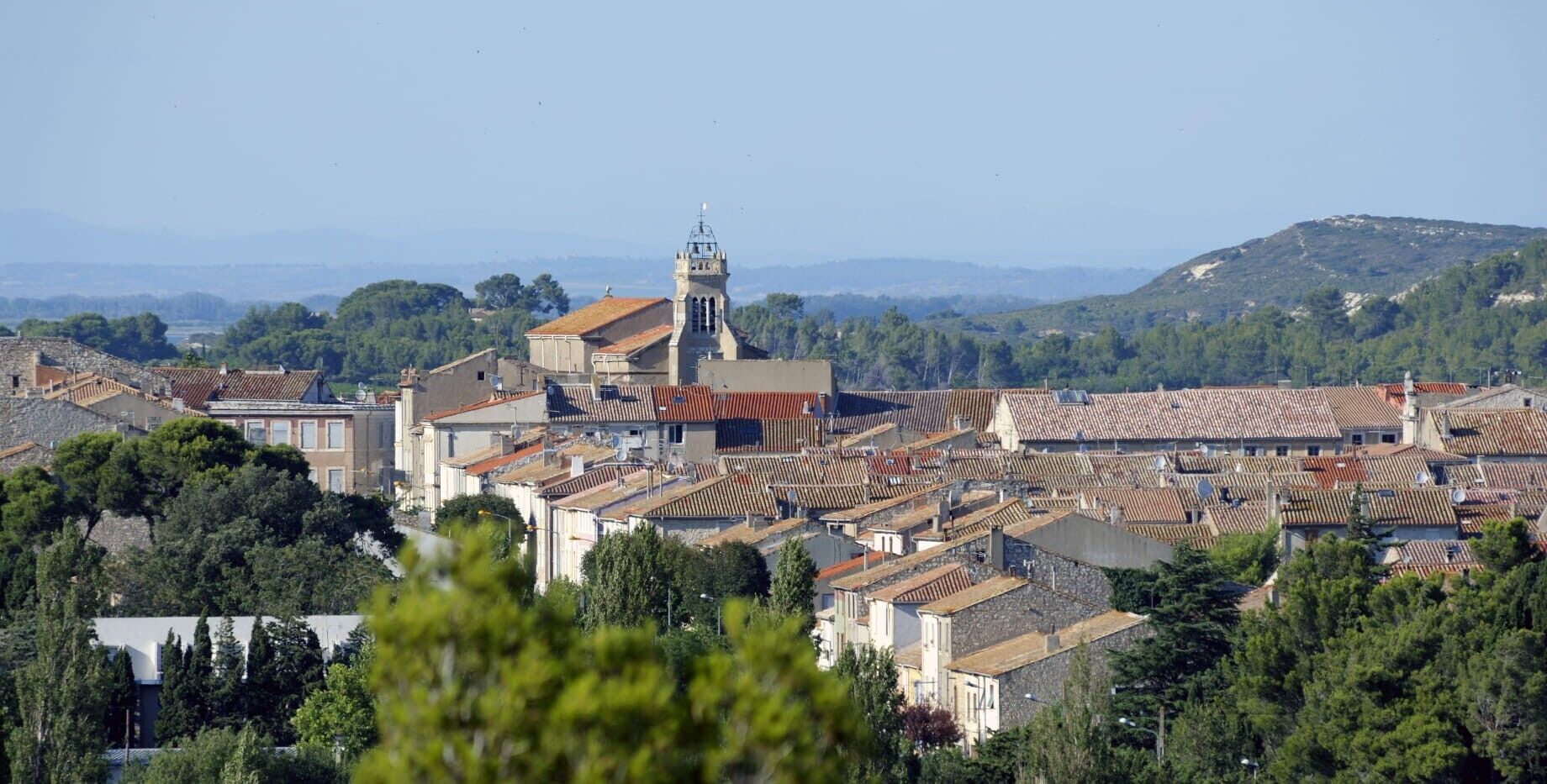 A town with terracotta-roofed buildings, a prominent church with a bell tower, and a hilly landscape in the background.