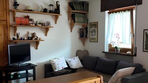 A living room with a wooden dining table, a sofa, a television, and wooden shelves with various items.