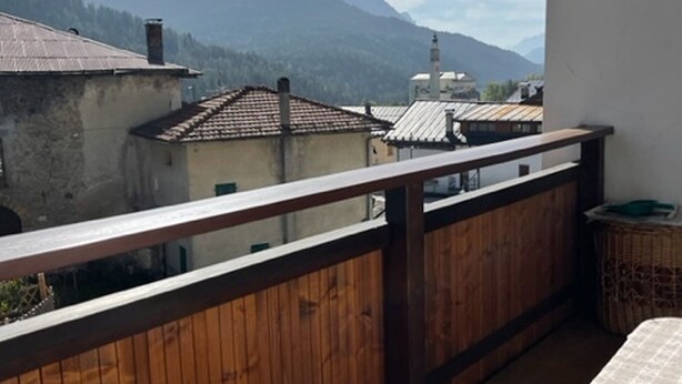 A balcony with a wooden railing, a table with a white tablecloth, and a view of buildings and mountains in the background.