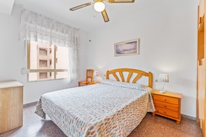 A bedroom with a bed, a wooden nightstand, a desk, a chair, a window with curtains, a ceiling fan, and a wall-mounted picture.