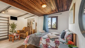 Un dormitorio con techo de madera, una cama con ropa de cama floral, un sofá, una mesa con una planta y una ventana con cortinas.