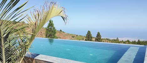A swimming pool with clear blue water, surrounded by stone tiles, and overlooking a coastal landscape with trees and a distant view of the sea.