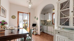 Een eethoek met een tafel, stoelen en een televisie. Een keuken met kasten en een raam.