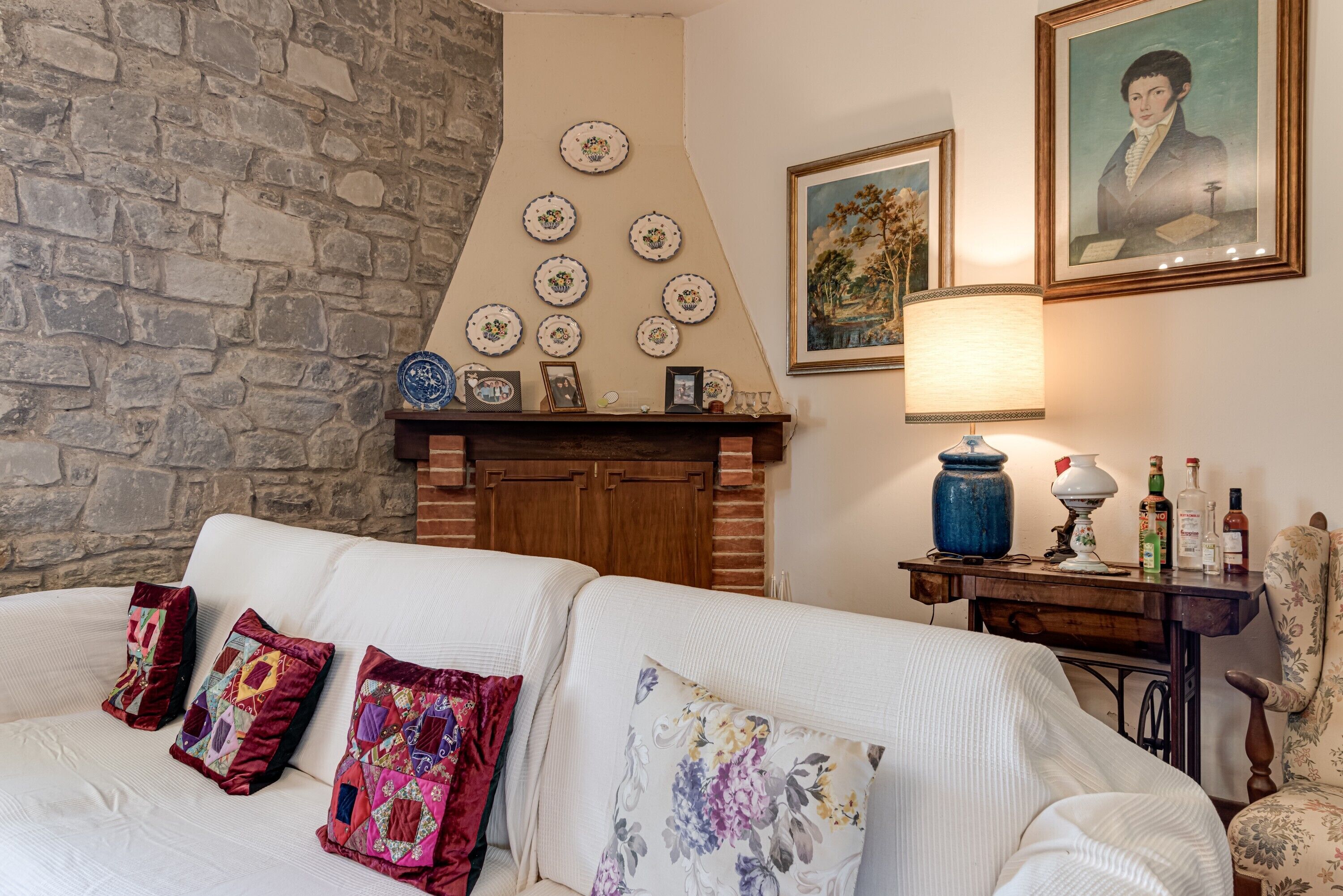 Une pièce avec un mur en pierre, une cheminée, un canapé blanc agrémenté de coussins colorés, une table en bois avec une lampe, et des œuvres d’art encadrées accrochées au mur.