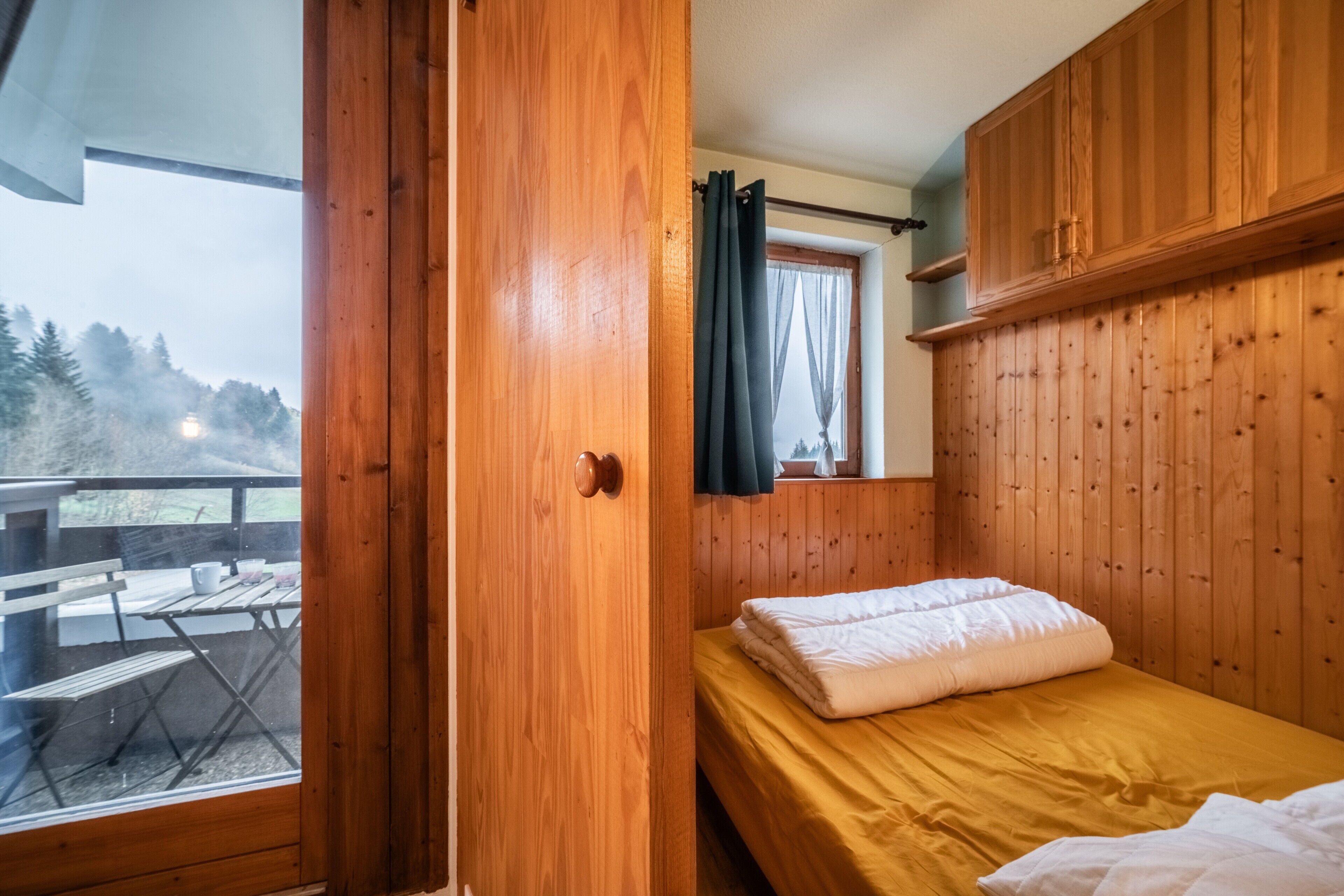 A wooden room with a bed, a window with curtains, and a view of a deck outside.